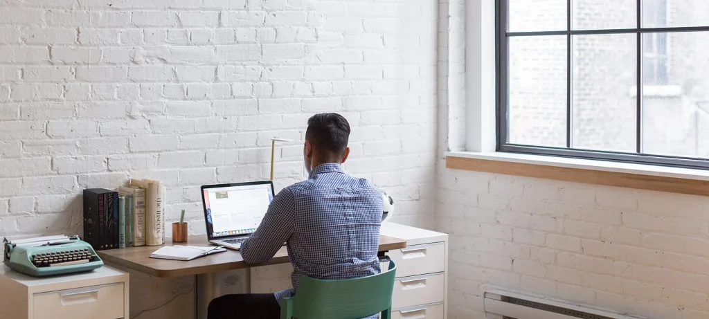 man working at a desk on his laptop facing a wall