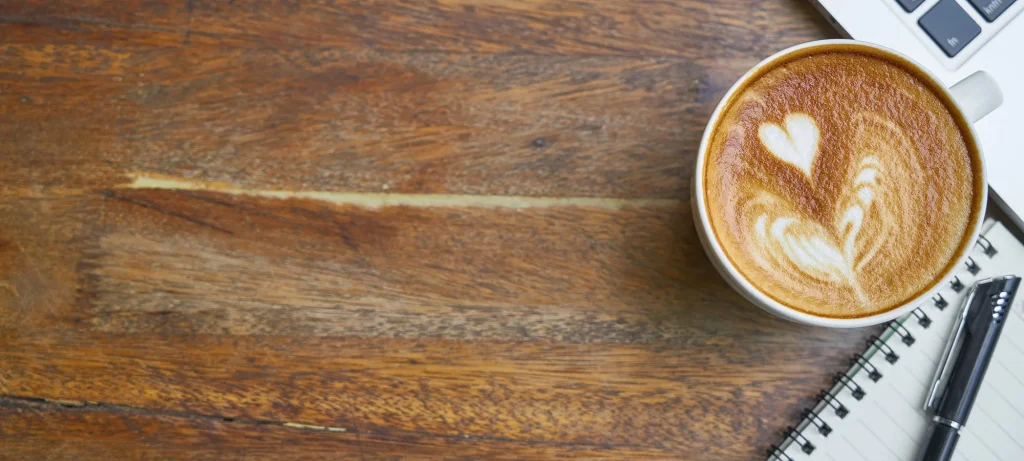 a close up of a cup of coffee on a wooden bench next to a laptop and notepad